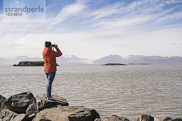 Junge Frau steht auf Felsblöcken und schaut durch ein Fernglas  Südost-Island