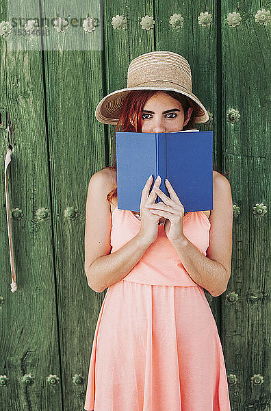 Porträt einer rothaarigen jungen Frau  die vor einer grünen Holztür steht und den Mund mit einem Buch bedeckt