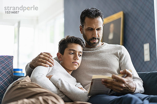 Vater liegt mit Sohn auf Couch im Wohnzimmer und liest Buch