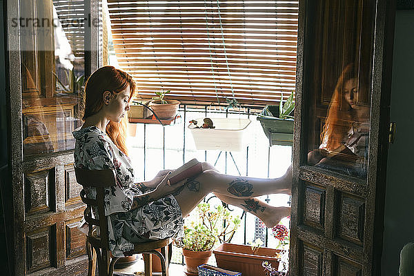 Rothaarige tätowierte Frau zu Hause beim Lesen eines Buches am Fenster