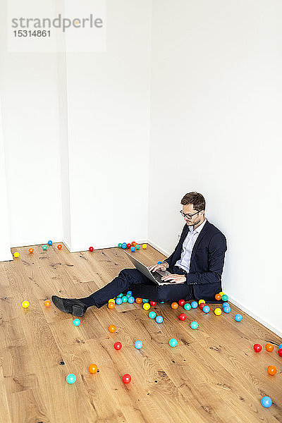 Auf dem Boden sitzender Geschäftsmann mit Laptop  umgeben von bunten Bällen