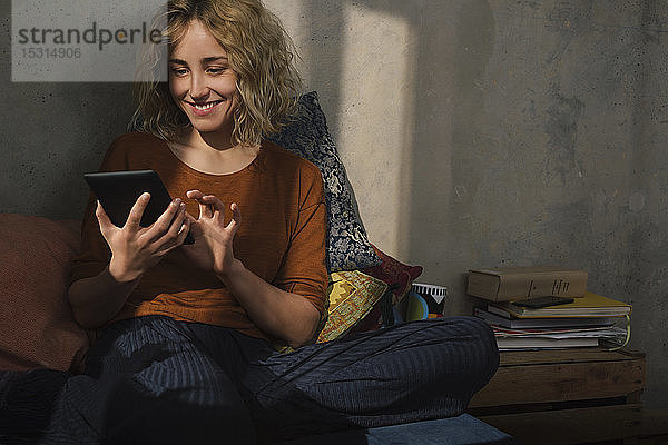 Porträt einer lächelnden jungen Frau  die mit einem E-Book-Reader auf dem Bett sitzt