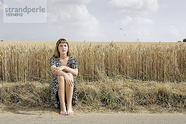 Porträt einer jungen Frau  die barfuss am Strassenrand vor einem Getreidefeld sitzt