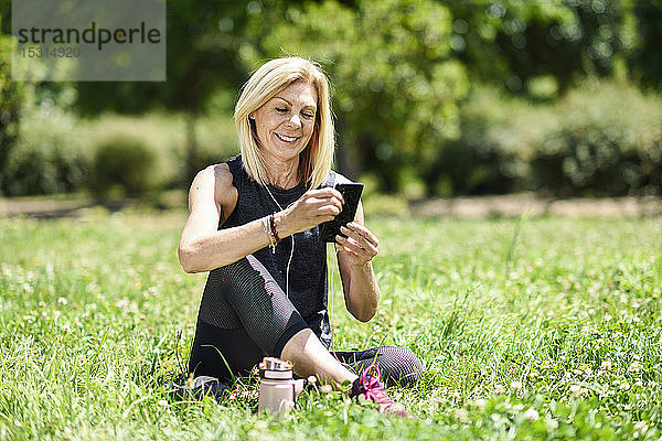 Reife Frau  die ihr Smartphone benutzt und sich nach dem Sport im Gras eines Parks ausruht