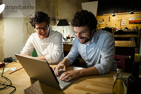 Zwei glückliche Kollegen benutzen gemeinsam einen Laptop in der Dunkelheit