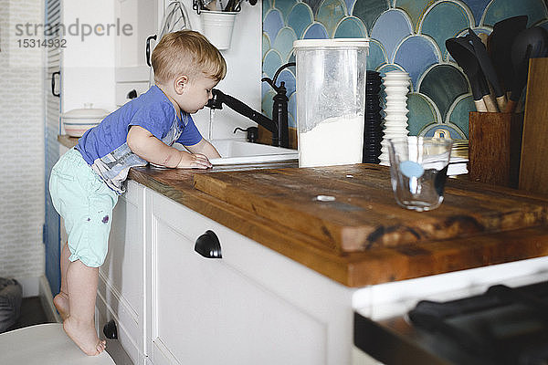 Kleiner Junge steht auf Zehenspitzen auf einem Stuhl in der Küche beim Geschirrspülen