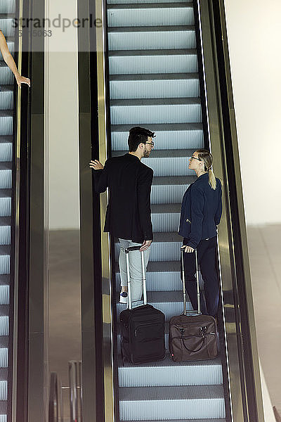 Zwei junge Geschäftspartner mit Gepäck unterhalten sich auf einer Rolltreppe