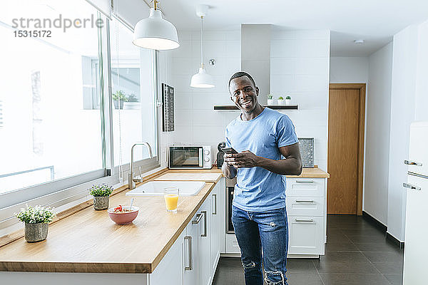 Porträt eines glücklichen jungen Mannes  der zu Hause in der Küche ein Handy benutzt