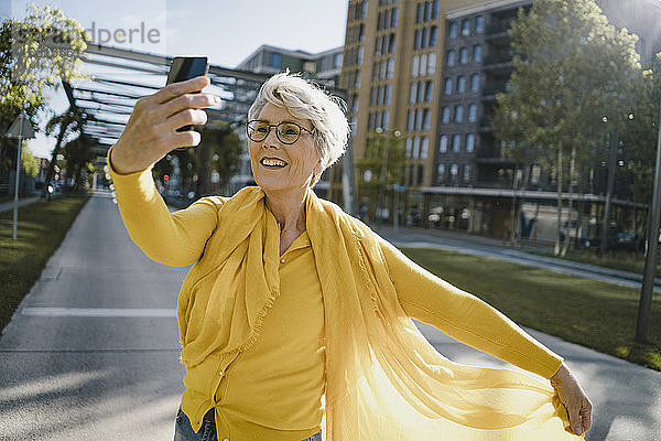 Porträt einer lächelnden reifen Frau in gelber Kleidung  die sich mit einem Smartphone selbstständig macht