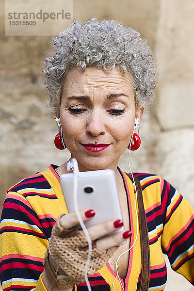 Porträt einer gepiercten reifen Frau mit grauem lockigem Haar beim Blick auf ein Mobiltelefon