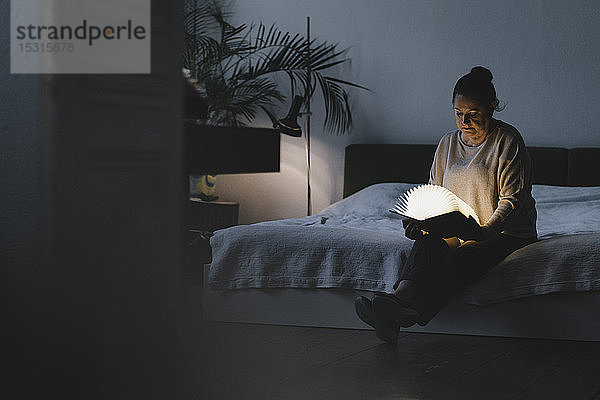 Ältere Frau sitzt auf Bett und hält leuchtendes Buch