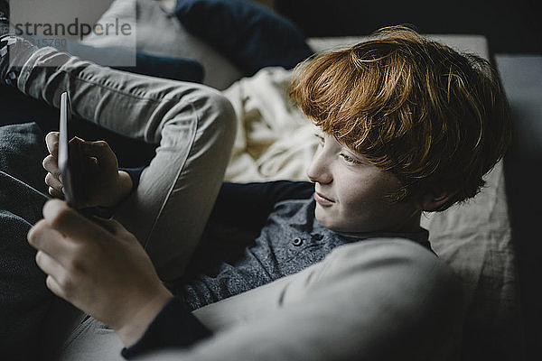 Rothaariger Junge liegt mit digitalem Tablett auf der Couch