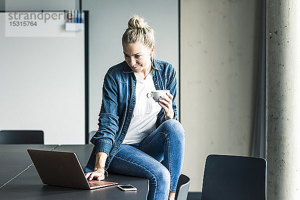 Geschäftsfrau mit Tasse Kaffee  die im Büro mit Laptop auf dem Tisch sitzt