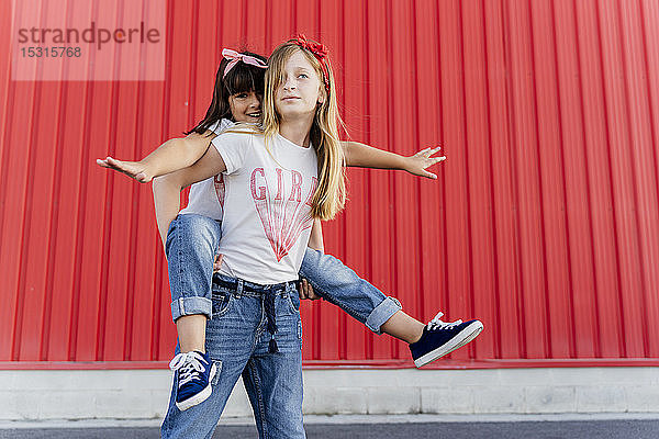 Mädchen trägt ihre Schwester auf dem Rücken und steht vor einer roten Wand