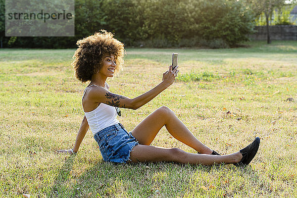 Tätowierte junge Frau  die auf einer Wiese sitzt und sich mit ihrem Smartphone selbstständig macht
