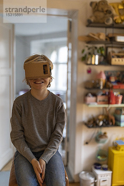 Junge sitzt auf einem Bock in seinem Zimmer zu Hause und trägt eine VR-Pappbrille