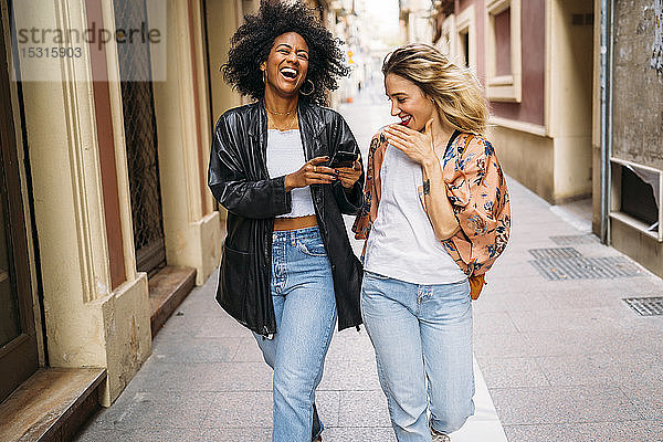 Multikulturelle glückliche Frauen  die in der Stadt spazieren gehen  auf Smartphones schauen und lachen