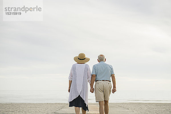 Rückenansicht eines älteren Ehepaares  das Hand in Hand auf einer Holzpromenade steht und aufs Meer blickt  Liepaja  Lettland