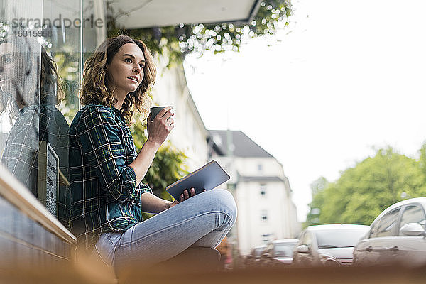 Junge Frau sitzt vor einem Café  benutzt ein digitales Tablet und trinkt Kaffee