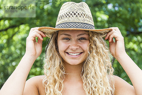 Porträt einer lächelnden blonden Frau mit Strohhut  Hände auf dem Hut