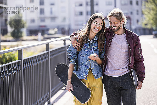 Glückliches junges Paar mit Skateboard und Smartphone auf einer Brücke