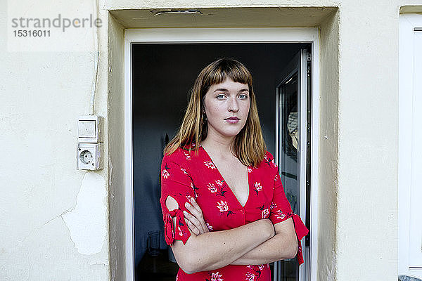 Porträt einer ernsten jungen Frau mit verschränkten Armen vor geöffneter Balkontür stehend