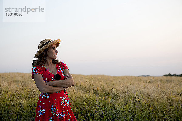 Frau mit Strohhut und rotem Sommerkleid mit Blumenmuster vor Getreidefeld stehend