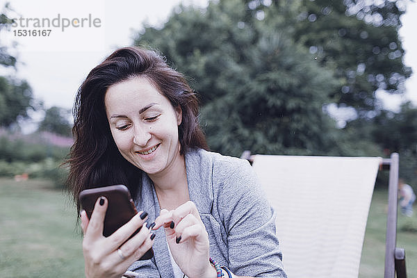 Lächelnde Frau benutzt Handy auf Liegestuhl im Garten