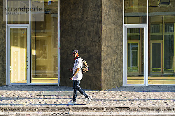 Junger Mann mit Rucksack geht bei Sonnenlicht auf dem Bürgersteig