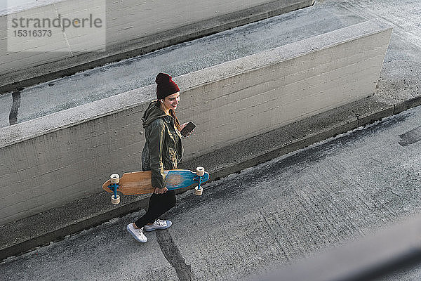 Stilvolle junge Frau mit Skateboard und Handy auf dem Parkdeck
