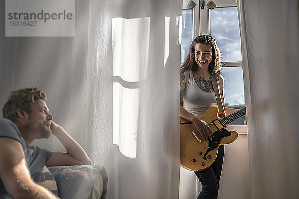 Mann schaut junge Frau zu Hause am Fenster mit einer Gitarre an