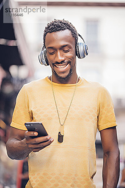Porträt eines lächelnden Mannes  der mit Kopfhörern Musik hört und auf ein Smartphone schaut