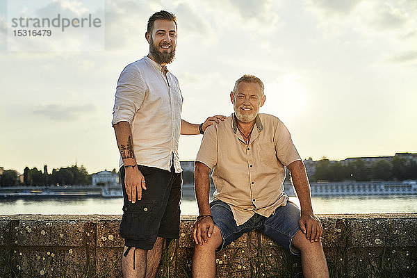 Porträt eines lächelnden Vaters und eines erwachsenen Sohnes am Flussufer bei Sonnenuntergang