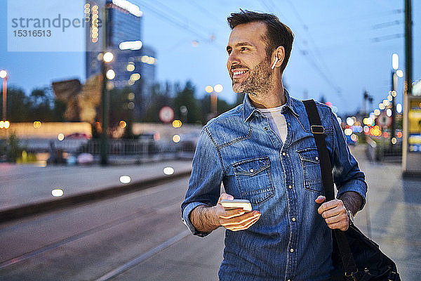 Lächelnder Mann benutzt Smartphone und hört Musik über drahtlose Kopfhörer  während er abends nach der Arbeit an der Straßenbahnhaltestelle wartet