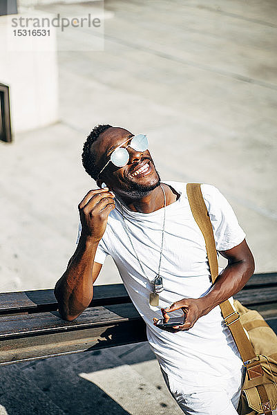 Glücklicher junger Mann mit Kopfhörern und Mobiltelefon in der Stadt