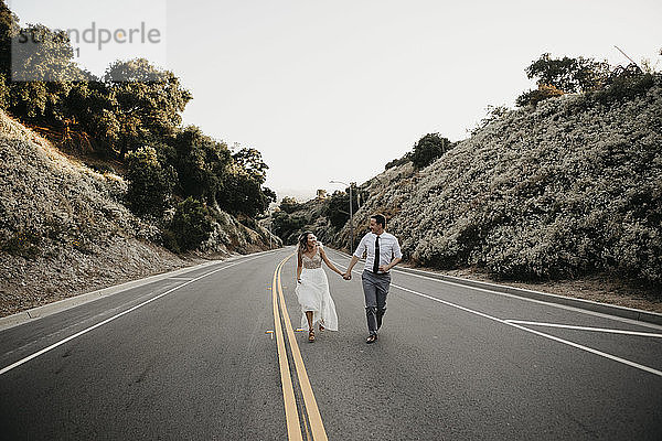 Braut und Bräutigam gehen Hand in Hand auf einer Landstraße