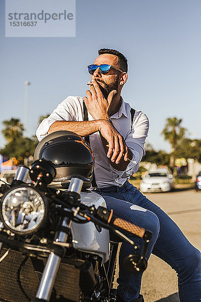Porträt eines bärtigen Motorradfahrers mit verspiegelter Sonnenbrille  der auf seinem Motorrad sitzt und raucht