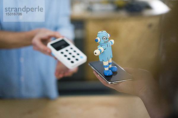 Kunde bezahlt berührungslos in einem Café  balanciert Spielzeugroboter auf Smartphone
