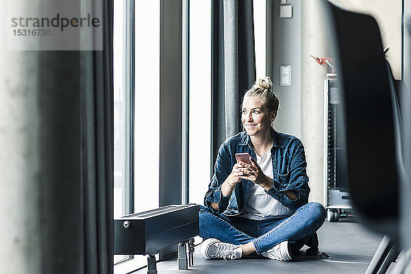 Lächelnde Geschäftsfrau sitzt im Büro mit Handy auf dem Boden