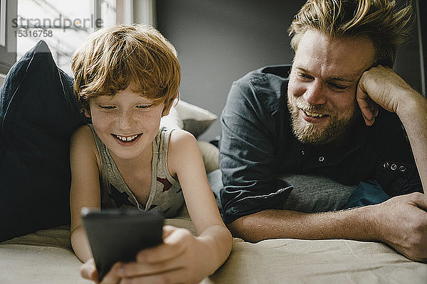 Porträt von glücklichem Vater und Sohn  die zusammen auf der Couch liegen und auf ihr Handy schauen