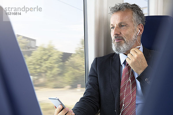 Älterer Geschäftsmann im Zug sitzend  mit Kopfhörern und Smartphone