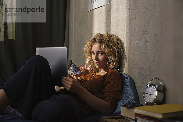 Porträt einer blonden jungen Frau  die mit einem Laptop auf dem Bett liegt