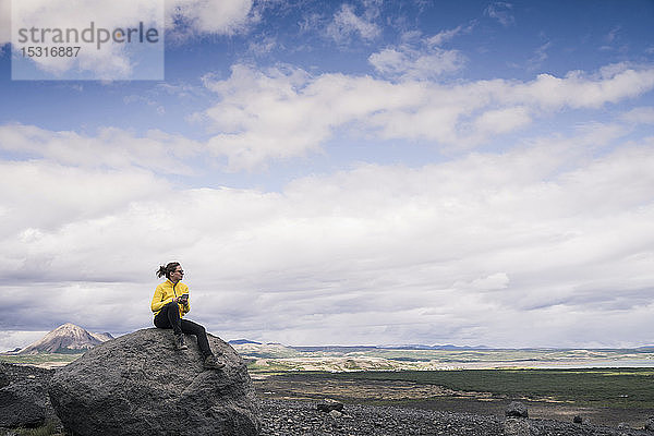 Junge Frau sitzt auf einem Felsen in Myvatn  Island  und benutzt ein Smartphone
