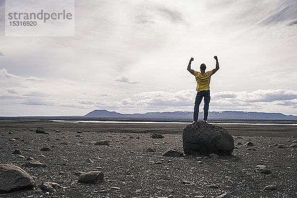 Erwachsener Mann steht mit erhobenen Armen auf einem Felsen im vulkanischen Hochland von Island