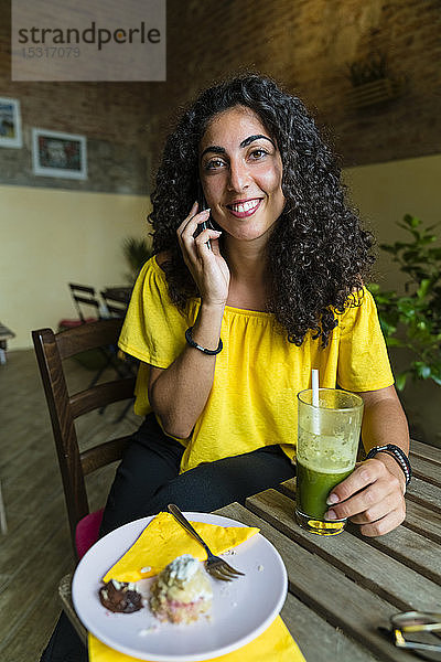 Porträt einer lächelnden jungen Frau am Handy  die mit einem Smoothie am Tisch sitzt