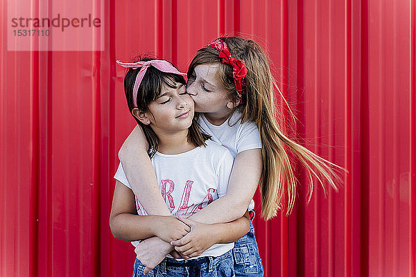 Zwei Schwestern stehen vor einer roten Wand und küssen sich