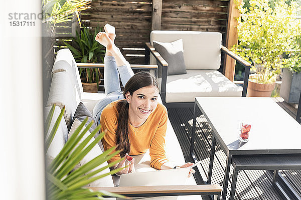 Porträt einer lächelnden jungen Frau  die mit Handy und Laptop auf der Couch auf der Terrasse liegt