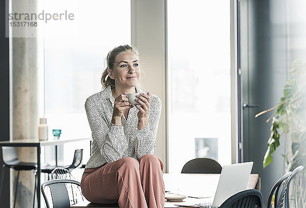 Lächelnde Geschäftsfrau sitzt im Büro am Tisch und macht eine Kaffeepause