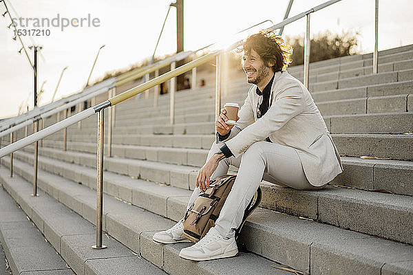 Lachender Geschäftsmann mit Kaffee zum Mitnehmen und Rucksack auf der Treppe im Freien sitzend