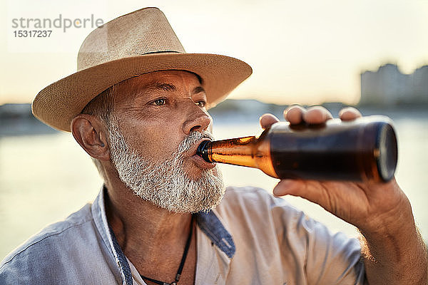 Älterer Mann trinkt bei Sonnenuntergang ein Bier am Flussufer